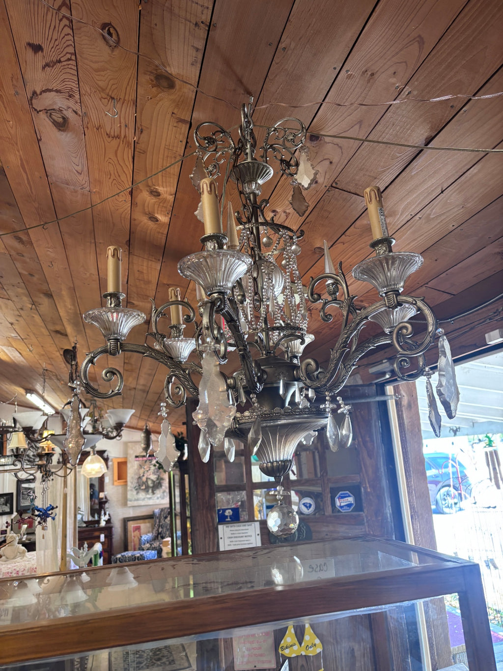 Vintage-style chandelier features an ornate silver-toned metal frame with intricate scrolling details, multiple arms holding light bulbs, and numerous hanging crystal prisms