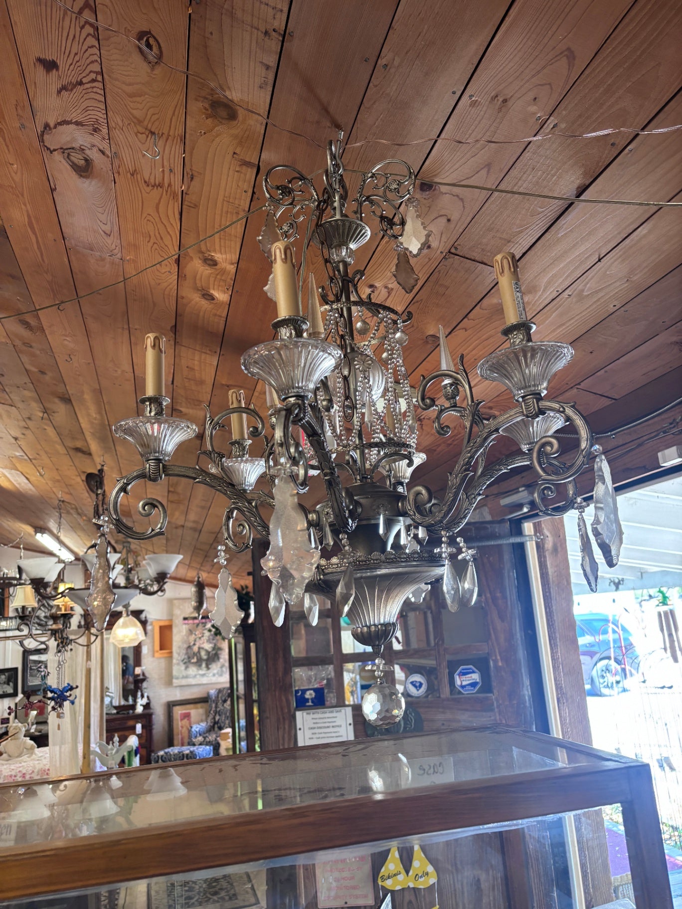 Vintage-style chandelier features an ornate silver-toned metal frame with intricate scrolling details, multiple arms holding light bulbs, and numerous hanging crystal prisms