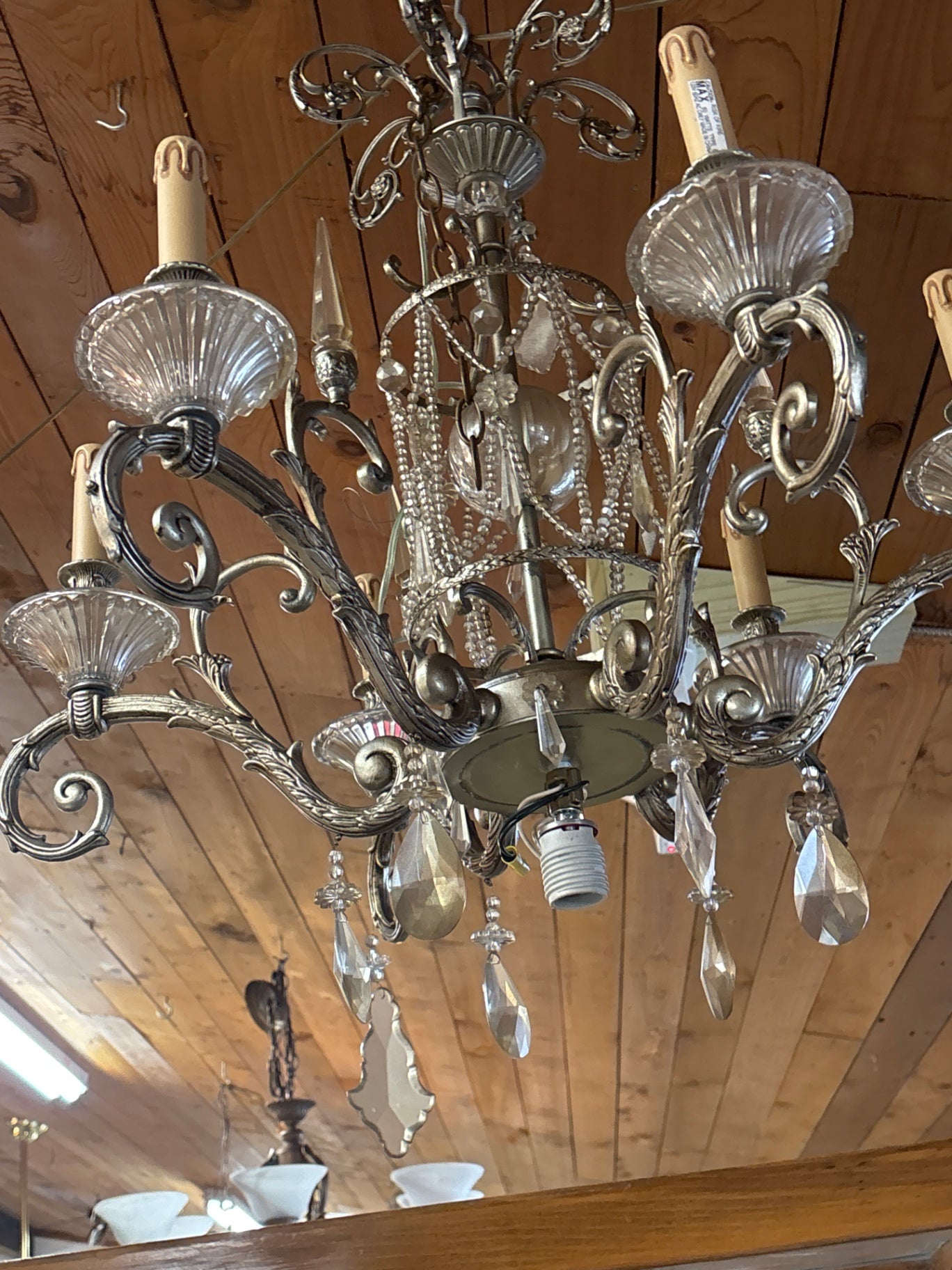 Vintage-style chandelier features an ornate silver-toned metal frame with intricate scrolling details, multiple arms holding light bulbs, and numerous hanging crystal prisms
