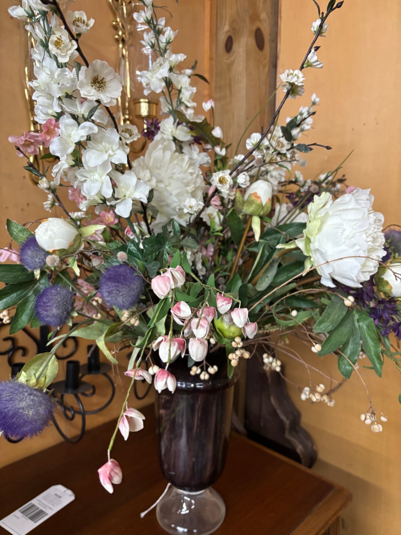This artificial floral arrangement features white peonies, white cherry blossoms, and purple thistle in a dark glass vase
