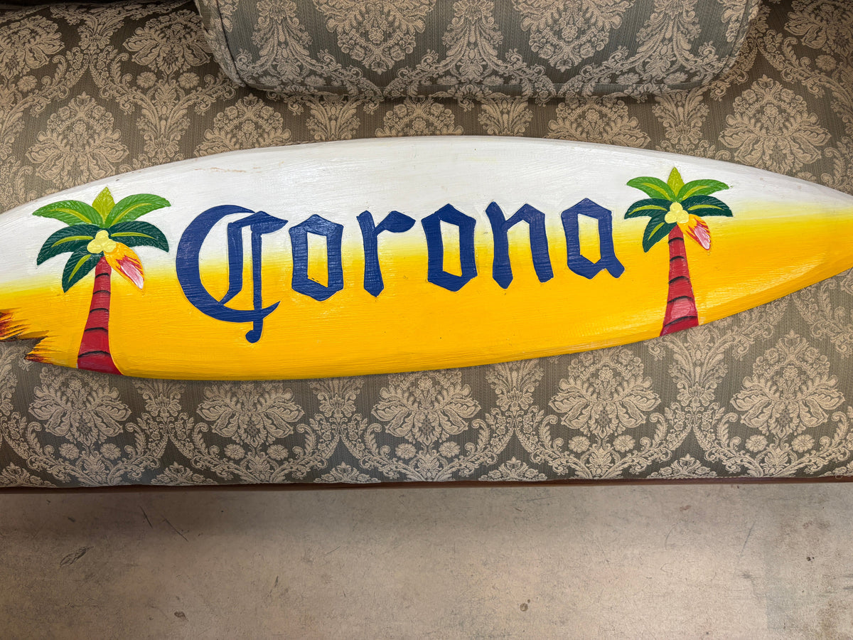 A hand-painted Corona surfboard featuring the brand name in blue lettering, yellow and white gradient sides, and painted palm trees, displayed on a patterned gray couch cushion.