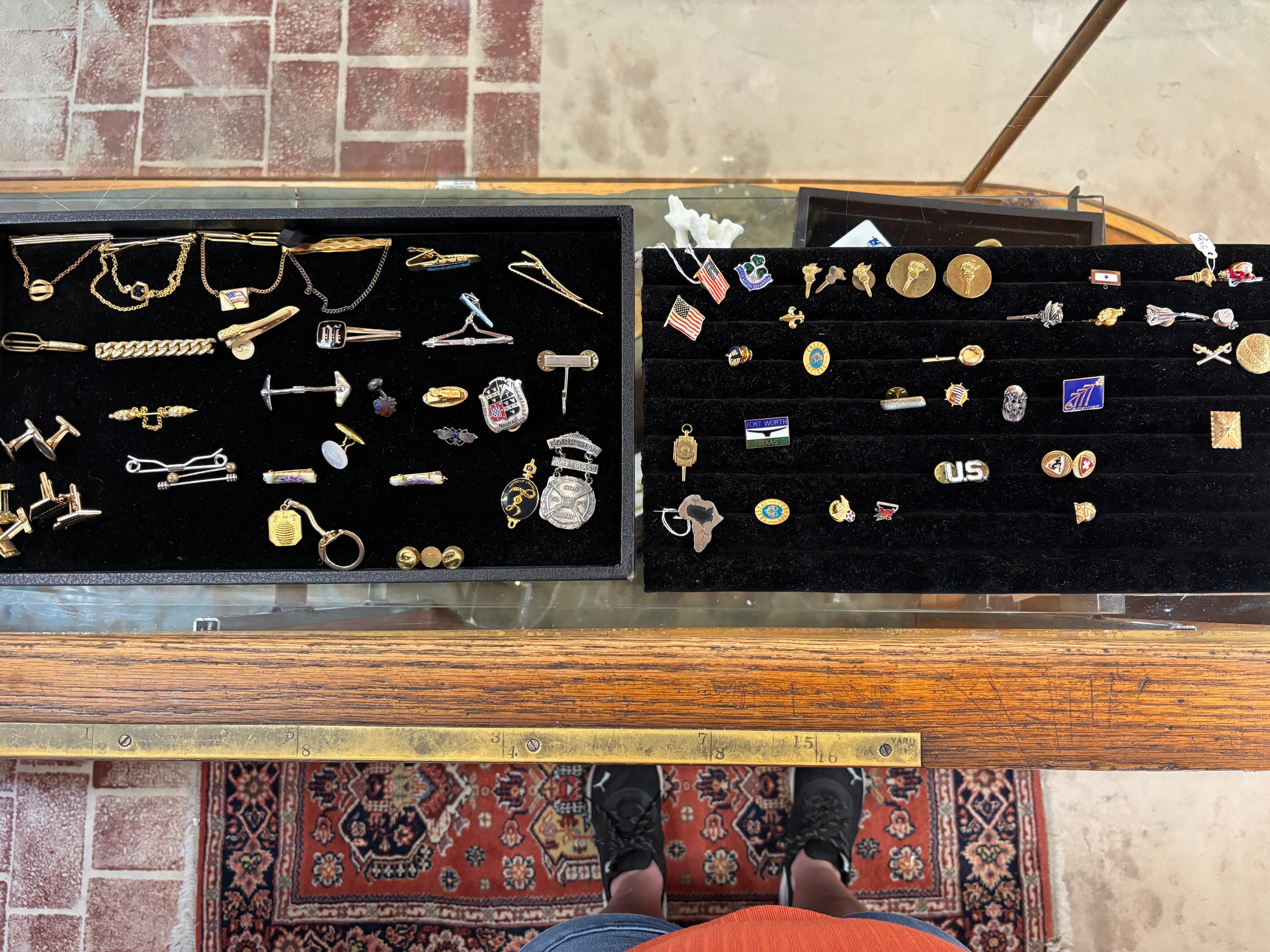 A display of assorted vintage and antique jewelry, primarily lapel pins and pins, arranged in two black velvet trays inside a wooden display case. 